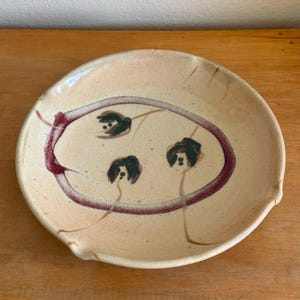 May include: A round ceramic plate with a speckled cream-colored glaze. The center features a hand-painted maroon oval border enclosing three dark green floral motifs with thin stems. The plate has a slightly irregular rim and a smooth finish.