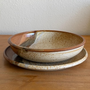 May include: A ceramic bowl and matching plate set featuring a speckled cream and tan glaze. The pieces have a smooth, rounded rim with a darker brown band along the edge, showcasing a natural, earthy aesthetic with a glossy finish throughout.
