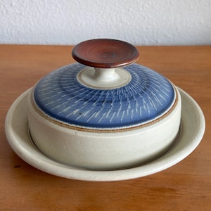 May include: A ceramic butter dish with a blue and white patterned lid and a small, round, brown handle. The dish sits on a matching cream-colored base, perfect for serving butter. The design is simple and elegant.