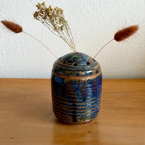 May include: A handmade ceramic vase with a blue and brown glaze. The vase has a rounded top with several holes and is filled with dried flowers and decorative grasses. The vase is sitting on a wooden surface.
