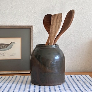 May include: A dark gray ceramic kitchen utensil holder with three wooden spoons. The holder is cylindrical with a slightly rounded top. A framed bird print is visible in the background.