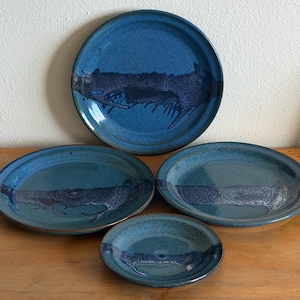 May include: A set of four blue ceramic plates with a dark blue abstract design. The plates vary in size, including a large dinner plate, two oval plates, and a small bowl. The plates are arranged on a wooden surface.