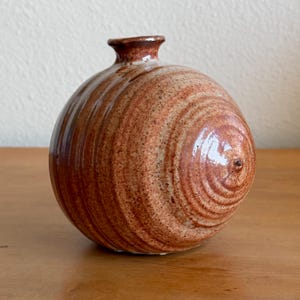 May include: A spherical ceramic vase with a narrow neck and a glossy finish. The surface features a speckled, earthy brown and tan glaze with concentric circular patterns radiating from the center, creating a textured appearance on the round body.