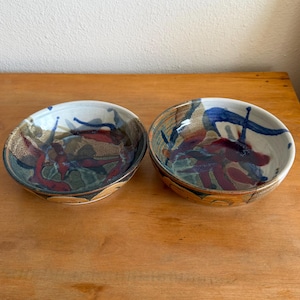 May include: Two handmade ceramic bowls with abstract designs. The bowls feature a cream-colored interior with blue, red, and brown brushstrokes. The exterior has a brown and gold pattern. The bowls are placed on a wooden surface.