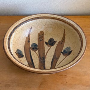 May include: A round ceramic bowl with a speckled cream-colored glaze and a dark brown rim. The interior features hand-painted brown vertical brushstrokes and dark blue floral motifs. The exterior base is a solid, smooth, light brown ceramic finish.