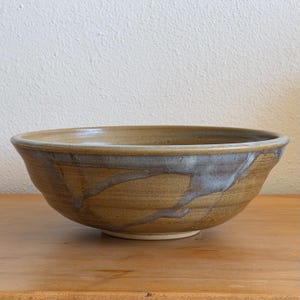 May include: A wide, round ceramic bowl featuring a warm, earthy tan base glaze. The exterior is decorated with a flowing, dripped pattern of cool, muted blue-grey glaze that cascades down the sides, creating a distinct contrast against the tan.