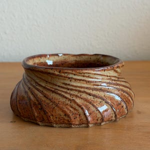 May include: A low, wide ceramic bowl with a glossy finish, featuring a spiraling, ribbed texture along the exterior. The glaze is a mottled blend of warm tan, brown, and cream tones with dark brown speckles throughout the entire surface.