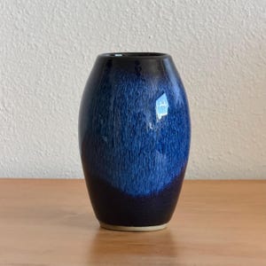 May include: A ceramic vase with a rounded, barrel-like shape featuring a glossy glaze. The surface displays a deep navy blue base color that transitions into a vibrant, streaked cobalt blue pattern toward the center, with a smooth rim.