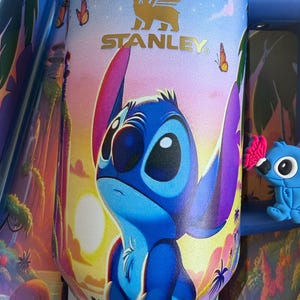 May include: A Stanley Flowstate tumbler featuring a colorful illustration of Stitch from Lilo & Stitch. The blue and purple character is set against a sunset background with butterflies. The tumbler has a gold Stanley logo and the text "THE FLOWSTATE".