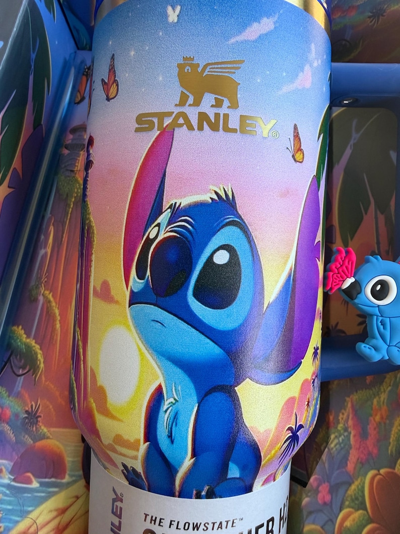 May include: A Stanley Flowstate tumbler featuring a colorful illustration of Stitch from Lilo & Stitch. The blue and purple character is set against a sunset background with butterflies. The tumbler has a gold Stanley logo and the text "THE FLOWSTATE".