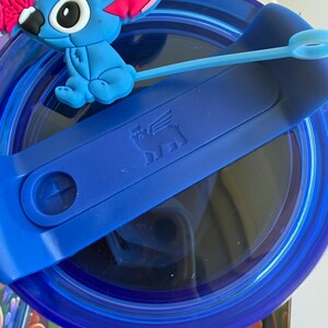 May include: A blue tumbler with a blue lid and a blue silicone straw. The lid features a blue Stitch character figurine with pink ears and a pink bow. The tumbler has a dark blue interior.