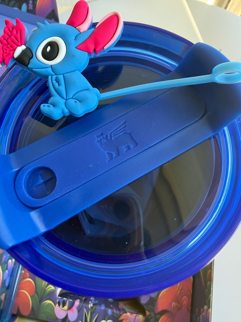 May include: A blue tumbler with a blue lid and a blue silicone straw. The lid features a blue Stitch character figurine with pink ears and a pink bow. The tumbler has a dark blue interior.