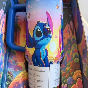 May include: A blue and white insulated travel mug featuring the character Stitch from Lilo & Stitch, set against a tropical sunset scene with butterflies. The mug has a blue handle and is in its original box.