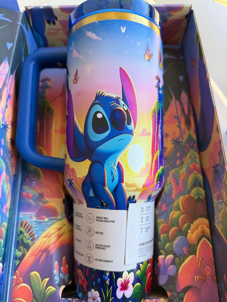 May include: A blue and white insulated travel mug featuring the character Stitch from Lilo & Stitch, set against a tropical sunset scene with butterflies. The mug has a blue handle and is in its original box.