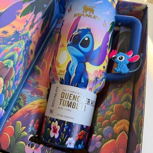 May include: A blue Stanley Quencher H2.0 Tumbler with a cartoon character design, featuring a blue character with large ears. The tumbler is 40 oz (1.18 L) and comes in a decorative box with a tropical landscape design. The text on the tumbler reads "All Day Hydration."