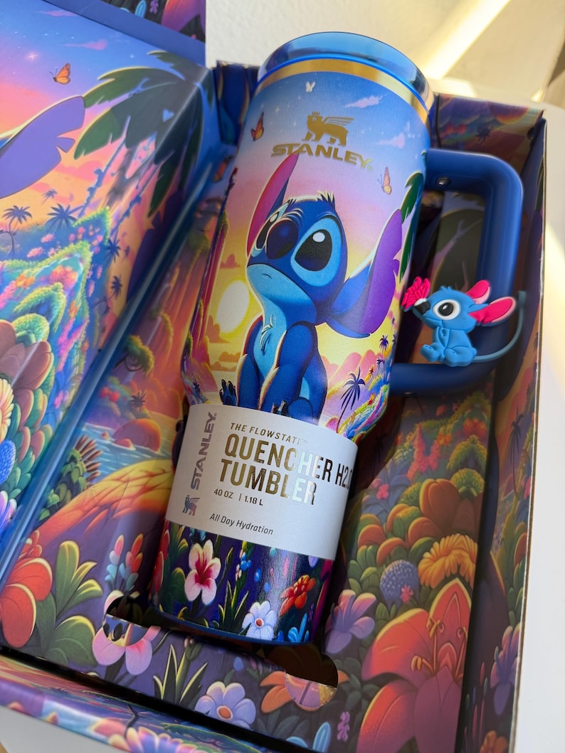 May include: A blue Stanley Quencher H2.0 Tumbler with a cartoon character design, featuring a blue character with large ears. The tumbler is 40 oz (1.18 L) and comes in a decorative box with a tropical landscape design. The text on the tumbler reads "All Day Hydration."