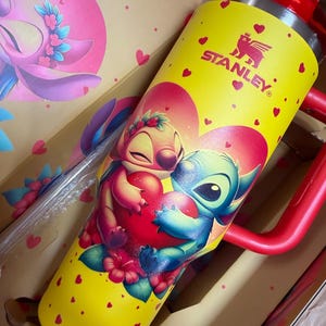 May include: A yellow Stanley tumbler featuring a graphic of Stitch and Angel hugging a red heart, surrounded by small red hearts. The tumbler has a red handle, a red lid, and a clear straw. The design includes floral accents and the Stanley logo.
