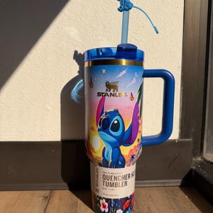 May include: A blue Stanley Quencher tumbler featuring a colorful design of the character Stitch from Lilo & Stitch. The tumbler has a blue handle, lid, and straw with a Stitch figure on top. The tumbler is 40 oz.