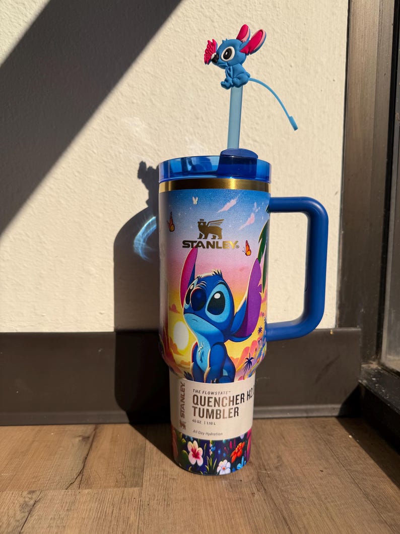 May include: A blue Stanley Quencher tumbler featuring a colorful design of the character Stitch from Lilo & Stitch. The tumbler has a blue handle, lid, and straw with a Stitch figure on top. The tumbler is 40 oz.