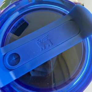 May include: A close-up of a blue tumbler lid with a handle. The lid is transparent with a blue rim and a blue handle. The handle has a small embossed image of a pig with wings. The lid is designed to fit a tumbler.