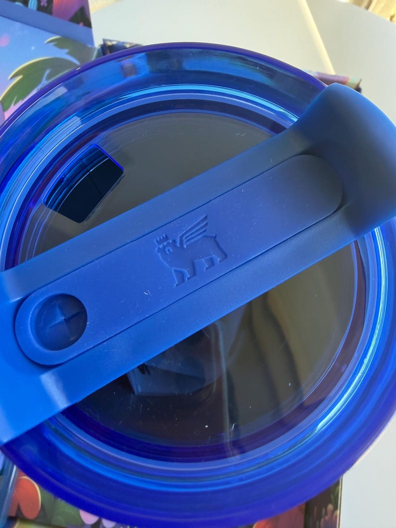 May include: A close-up of a blue tumbler lid with a handle. The lid is transparent with a blue rim and a blue handle. The handle has a small embossed image of a pig with wings. The lid is designed to fit a tumbler.