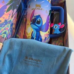 May include: A blue Stanley mug featuring the character Stitch from Lilo & Stitch, with a matching blue handle and a small Stitch figure attached. The mug is in a decorative box with a blue cloth and the Stanley and Loveshackfancy logos.