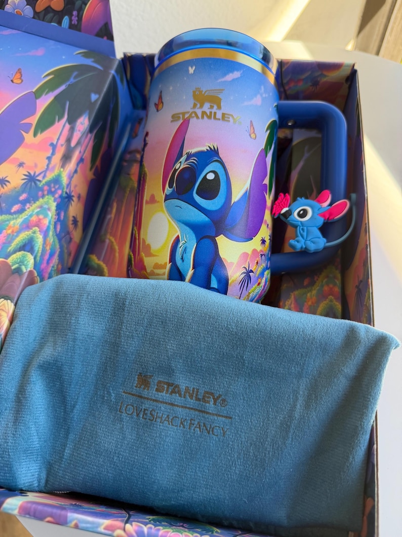 May include: A blue Stanley mug featuring the character Stitch from Lilo & Stitch, with a matching blue handle and a small Stitch figure attached. The mug is in a decorative box with a blue cloth and the Stanley and Loveshackfancy logos.