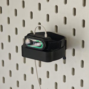 Op de afbeelding: Een zwarte smartwatch die oplaadt op een zwart oplaadstation. Het horloge geeft de tijd weer, 12:55, en een batterijniveau van 65%. Een witte oplaadkabel is aangesloten op het horloge en het station. Het station is gemonteerd op een wit pegboard.