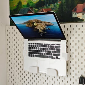 May include: A white laptop computer is mounted on a white pegboard using white brackets. The laptop screen displays a scenic image of an island in the ocean. The pegboard has evenly spaced holes.
