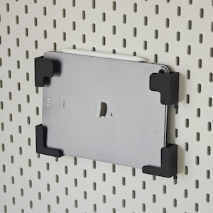 Puede incluir: Un iPad plateado con un lápiz óptico blanco está montado en una pared perforada blanca con un soporte negro. El soporte tiene un diseño cuadrado y está fijado a la pared perforada con tornillos. El iPad está centrado en el marco.