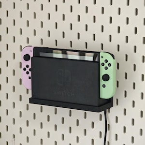 Puede incluir: Una consola Nintendo Switch negra con un Joy-Con rosa a la izquierda y un Joy-Con verde a la derecha. La consola está montada en un estante negro contra una pared de tablero perforado blanco. Un cable de carga negro está enchufado.