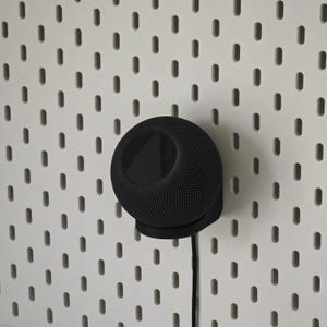 Op de afbeelding: Een zwarte, bolvormige luidspreker gemonteerd op een wit pegboard. De luidspreker heeft een mesh-achtige textuur en een cirkelvormige basis. Een zwarte kabel loopt naar beneden vanaf de luidspreker.