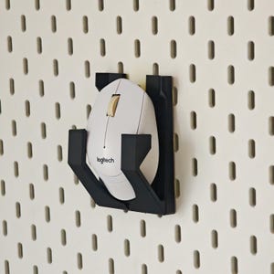 May include: A white Logitech computer mouse rests inside a black plastic wall-mounted holder. The holder is attached to a white pegboard with vertical slots. The mouse features a central scroll wheel and the Logitech brand name printed in black.