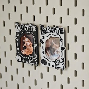 May include: Two cat-themed photo frames, one black and one white, each with a picture of a cat. The word "CUTE" is at the top of each frame. The frames have cat paw prints, fish, and heart designs.
