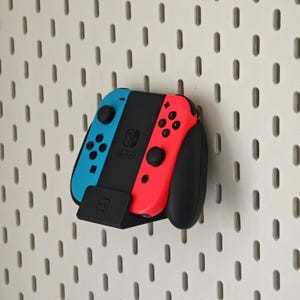 May include: A Nintendo Switch console with blue and red Joy-Con controllers is mounted on a white pegboard. The console is black with the Nintendo Switch logo. A black stand holds the controllers.