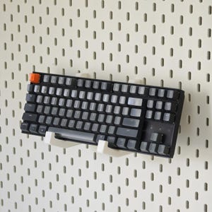 May include: A black mechanical keyboard with translucent gray keycaps and a single orange escape key, mounted on a white wall-mounted pegboard using two white plastic support brackets that hold the keyboard in a horizontal position.