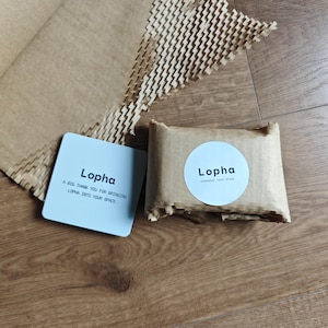 K&ouml;nnte beinhalten: Eine quadratische wei&szlig;e Karte mit dem schwarzen Text Lopha und einer Dankesnachricht, neben einem kleinen rechteckigen Paket, das in braunes Papier eingewickelt ist, mit einem runden wei&szlig;en Aufkleber mit dem schwarzen Text Lopha und Organize Your Space.