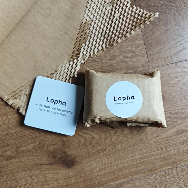 K&ouml;nnte beinhalten: Eine quadratische wei&szlig;e Karte mit dem schwarzen Text Lopha und einer Dankesnachricht, neben einem kleinen rechteckigen Paket, das in braunes Papier eingewickelt ist, mit einem runden wei&szlig;en Aufkleber mit dem schwarzen Text Lopha und Organize Your Space.
