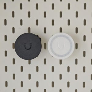 K&ouml;nnte beinhalten: Zwei kreisf&ouml;rmige Lochwandhaken, einer in Schwarz und einer in Wei&szlig;, jeweils mit einem mittig gepr&auml;gten Smiley-Symbol. Diese funktionalen Zubeh&ouml;rteile sind f&uuml;r die sichere Befestigung an einer wei&szlig;en Standard-Lochwand mit vertikalen Schlitzen.