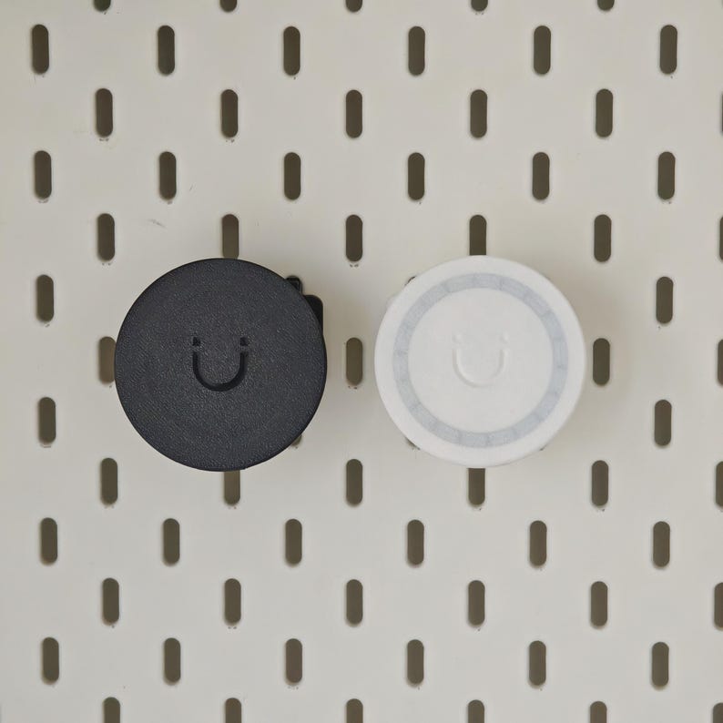 K&ouml;nnte beinhalten: Zwei kreisf&ouml;rmige Lochwandhaken, einer in Schwarz und einer in Wei&szlig;, jeweils mit einem mittig gepr&auml;gten Smiley-Symbol. Diese funktionalen Zubeh&ouml;rteile sind f&uuml;r die sichere Befestigung an einer wei&szlig;en Standard-Lochwand mit vertikalen Schlitzen.