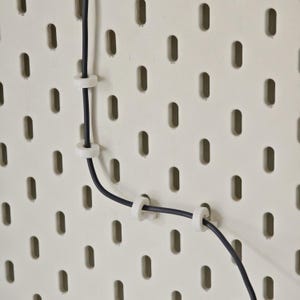 May include: Close-up of a cream-colored pegboard with evenly spaced oval holes. A black cable is secured to the board with white cable clips. The cable curves across the board, held in place by the clips.