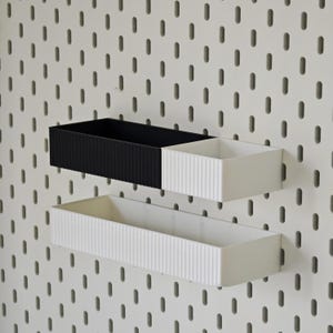 May include: Two rectangular wall-mounted storage containers. One is black with vertical ridges, the other is white with vertical ridges. A third white container is below. All are mounted on a white pegboard.