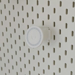 K&ouml;nnte beinhalten: Ein wei&szlig;er, runder Lochwandhaken mit einer flachen Vorderseite, die ein dezentes, gepr&auml;gtes Smiley-Symbol und einen d&uuml;nnen kreisf&ouml;rmigen Rand aufweist. Der Haken ist in eine wei&szlig;e Lochwand mit einem Muster aus vertikalen Schlitzen eingesetzt.