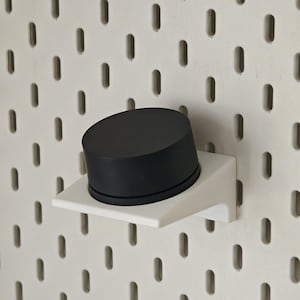 May include: A black, cylindrical object rests on a white, angled shelf. The shelf is mounted on a white pegboard with evenly spaced, vertical slots. The object appears to be a storage container or decorative item.