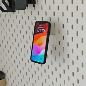 May include: A black smartphone with a colorful display is mounted on a white pegboard using a white holder. The phone screen displays the time as 01:19 on Sunday, January 25. The pegboard has a series of evenly spaced oval holes.
