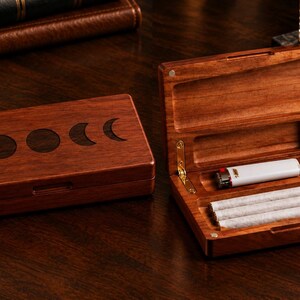 Peut inclure: Un étui rectangulaire en bois avec une incrustation de phases de lune sur le couvercle. L'étui ouvert révèle un briquet blanc et trois articles roulés rangés dans des compartiments dédiés, avec des charnières en métal doré et une finition polie.