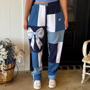 May include: Patchwork sweatpants featuring a color-blocked design in shades of navy blue, light blue, and white. The left leg displays a large white bow graphic with a blue floral pattern. A small white Nike logo is embroidered on the upper right thigh.