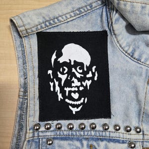 May include: A light blue acid-wash denim waistcoat featuring a rectangular black fabric patch sewn onto the chest. The patch displays a white, high-contrast graphic of a stylised, melting human face. A row of silver metal studs lines the lower seam.