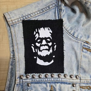 May include: A rectangular black fabric patch featuring a white screen-printed graphic of Frankenstein's monster, stitched onto a light blue acid-wash denim vest adorned with a horizontal row of silver-toned metal studs along the lower seam.