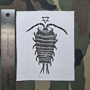 Pode incluir: Um emblema de tecido branco com uma ilustração a tinta preta de um isópode com um símbolo de triângulo acima da cabeça. Uma régua de aço inoxidável está posicionada à esquerda, mostrando que o emblema mede cerca de 11,5 cm de altura.